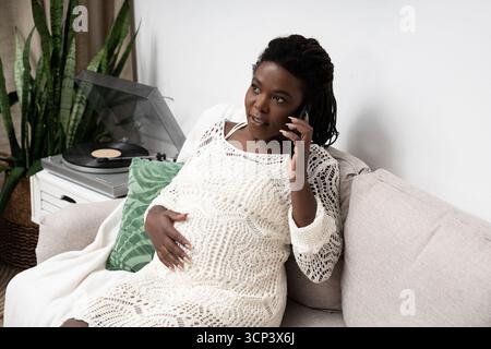 Une femme afro-américaine enceinte se détend sur son canapé, parlant au téléphone à la maison. Banque D'Images