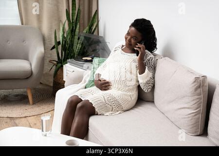 Une femme enceinte est assise confortablement sur le canapé, discutant au téléphone à la maison, embrassant la maternité. Banque D'Images