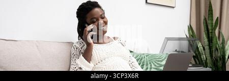 Une belle femme afro-américaine se détend à la maison tout en parlant au téléphone, bannière Banque D'Images