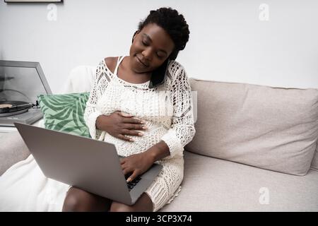 Une femme afro-américaine enceinte se détend à la maison, en utilisant son ordinateur portable avec les mains sur son ventre. Banque D'Images