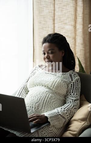 Femme enceinte se détend à la maison, travaillant sur son ordinateur portable, embrassant la maternité et le confort. Banque D'Images