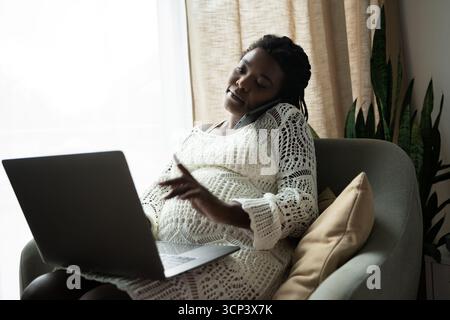 Une femme enceinte se détend à la maison tout en étant au téléphone et en utilisant son ordinateur portable. Banque D'Images