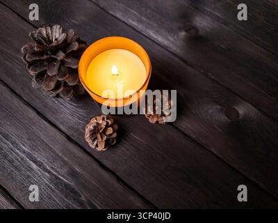 Un plat minimaliste avec une bougie en verre brun, des pommes de pin et une plume sur une surface en bois sombre. Idéal pour saisonnier, bien-être, ou intérieur de Banque D'Images