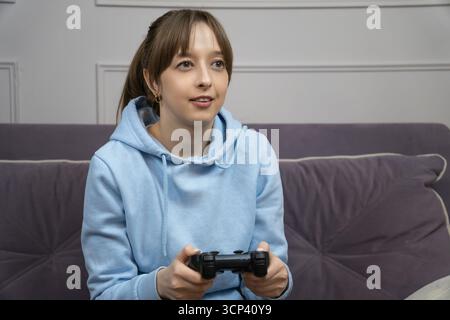 Gamer Girl assis confortablement sur un canapé confortable, tenant une manette et se plongeant dans des jeux vidéo passionnants, profitant du temps de loisirs à la maison et e Banque D'Images