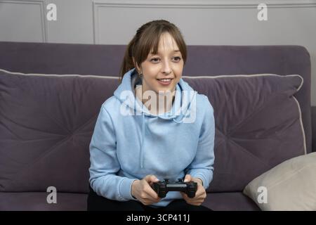 Gamer Girl assis confortablement sur un canapé confortable, tenant une manette et se plongeant dans des jeux vidéo passionnants, profitant du temps de loisirs à la maison et e Banque D'Images