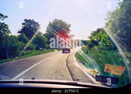 Photo analogique prise depuis le siège passager, montrant une Ford d'époque conduisant devant vous. Scène Road trip avec ambiance rétro et mouvement. Banque D'Images