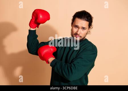 Jeune homme posant en toute confiance avec des gants de boxe rouges sur un fond beige neutre, portant un pull vert élégant Banque D'Images