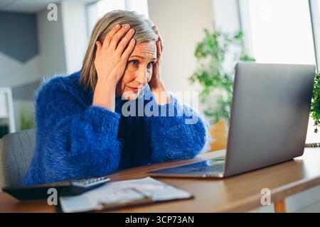 Femme âgée portant un pull bleu semblant stressé tout en utilisant un ordinateur portable dans un cadre de maison confortable pendant la journée Banque D'Images