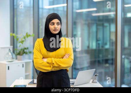 Femme d'affaires musulmane portant un hijab et une chemise jaune debout avec confiance avec les bras pliés dans un cadre de bureau contemporain, représentant la réussite professionnelle et la détermination Banque D'Images