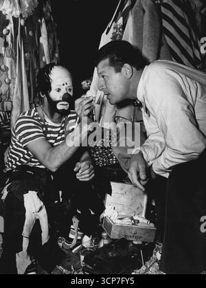Enfilant la garde-robe de Clown qu'il porte dans sa dernière sortie, « April Showers ». Jack Carson s'est joint à une conspiration avec les interprètes du Clyde Beauty Circus pour susciter une agréable surprise sur le public lors de l'ouverture du spectacle à Los Angeles. Jouer toute la performance incognito, The Warner Bros Star a suscité des cris de joie lorsqu'il a révélé sa véritable identité lors de la finale de la série. Clown Mark Anthony vérifie un faux nez pour la taille alors que le comédien Warner se prépare à revêtir le maquillage et la garde-robe pour son apparition sous la grande tente. 11 novembre 1948. (Photo de Floyd McCarty) Banque D'Images