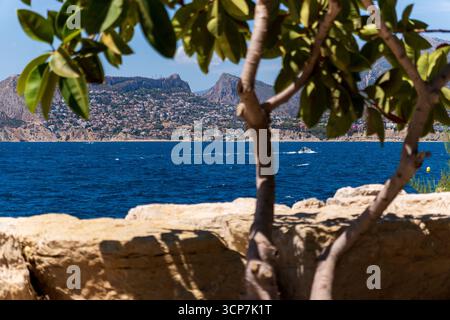 Scène estivale : des branches d'arbres encadrent élégamment un bateau à moteur sur la mer bleue, avec les montagnes emblématiques de Calpe en arrière-plan. Banque D'Images