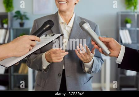 Parlant avec confiance en souriant, femme dirigeante interviewée au bureau par des journalistes tenant des microphones Banque D'Images