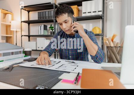 Jeune architecte confiant travaillant au bureau, dessinant des plans de maison et discutant des détails de construction au téléphone, en se concentrant sur la précision et la création Banque D'Images
