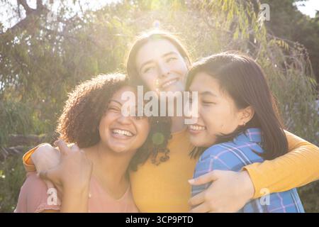 Diverses amies féminines debout dans le parc serrant les épaules vers la lumière du soleil filtrant à travers les arbres Banque D'Images