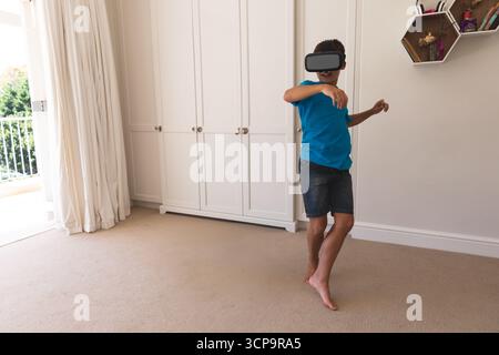 Garçon d'âge scolaire portant un T-shirt bleu explorant VR à l'aide d'un casque dans la chambre près des étagères, espace de copie Banque D'Images