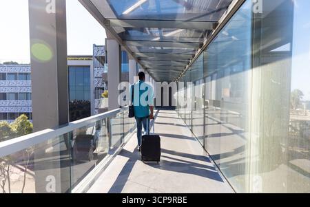 Homme afro-américain marchant à travers le couloir de verre tirant la valise et portant le sac à bandoulière Banque D'Images