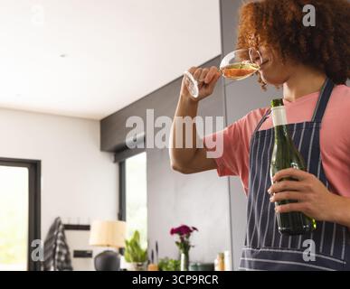 Femme adulte afro-américaine dans un tablier tenant une bouteille de vin sirotant du verre dans la cuisine, espace de copie Banque D'Images