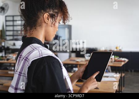 Femme adulte moyenne tapant la tablette tout en drapant le torchon par des tables en bois dans le restaurant, espace de copie Banque D'Images
