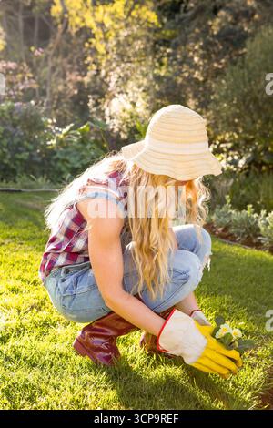Jardinière féminine agenouillée dans le jardin de l'arrière-cour tenant des fleurs jaunes portant des gants de jardin espace de copie Banque D'Images