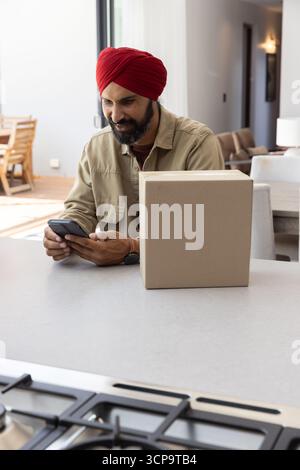 Homme indien portant turban rouge à l'aide d'un smartphone assis à l'îlot de cuisine déballage cardboa Banque D'Images