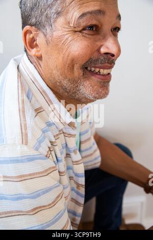 Homme senior souriant tout en étant assis contre un mur pâle portant une chemise à rayures pastel, espace copie Banque D'Images