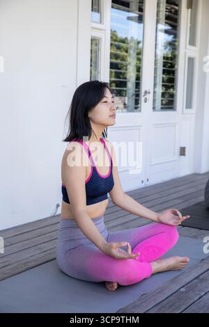 Femme asiatique méditant les jambes croisées sur le tapis de yoga gris sur le pont du porche en bois portant des vêtements de sport Banque D'Images