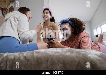 Diverses amies féminines se prélassant dans la chambre à coucher en appliquant des masques faciaux avec brosse et tenant smartphone Banque D'Images