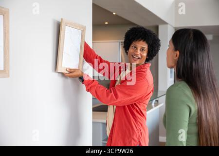 Couple diversifié accrochant des œuvres d'art encadrées en bois sur le mur de couloir de la maison près du comptoir Banque D'Images