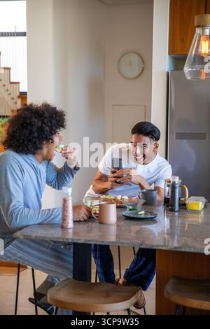 Divers amis masculins mangeant des toasts d'avocat et sirotant du café à l'îlot de cuisine avec smartphone Banque D'Images