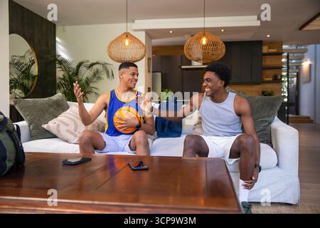Divers amis masculins assis sur le canapé haut-fiving, tenant basket-ball et smartphone dans le salon Banque D'Images