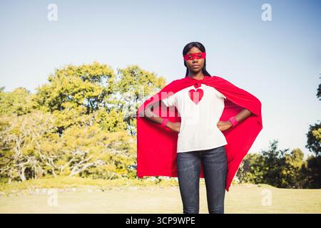 Femme afro-américaine posant dans le parc portant une cape de super-héros rouge, un masque, des bracelets et un tee-shirt coeur Banque D'Images