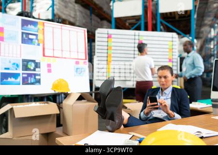 Gestionnaire féminin assis sur le bureau à l'aide d'un smartphone parmi les boîtes, tableaux dans l'entrepôt, espace de copie Banque D'Images