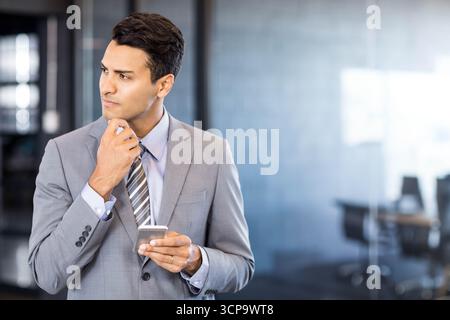 Professionnel d'affaires tenant smartphone et touchant menton soigneusement dans le bureau avec des murs de verre Banque D'Images