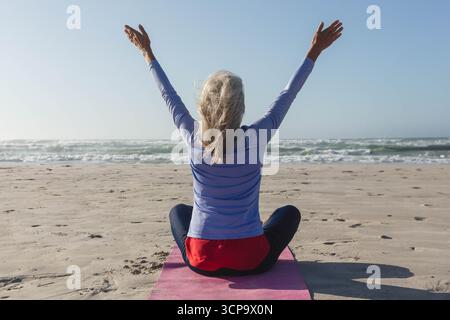 Femme senior pratiquant le yoga assise croisée sur un tapis rose au bord de l'océan portant des vêtements de sport Banque D'Images
