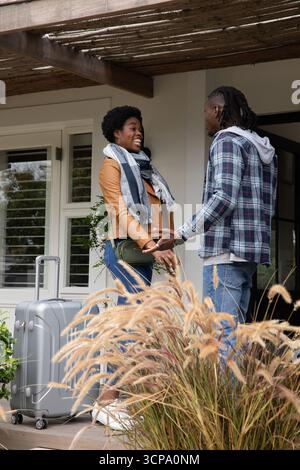 Couple afro-américain tenant la main et saluant sur le porche couvert de pergola avec valise Banque D'Images