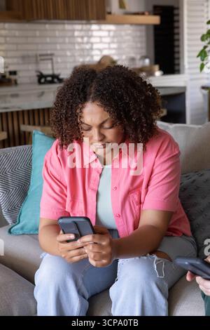 Amis afro-américains assis sur un canapé gris avec oreiller sarcelle dans la cuisine ouverte à l'aide de smartphones Banque D'Images