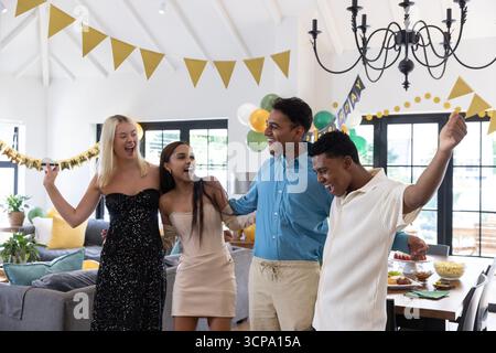 Divers amis debout sous des bannières de fanion d'or avec des ballons dans le salon Banque D'Images