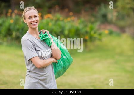 Bénévole souriante transportant un sac en plastique vert dans un parc public portant un t-shirt gris de bénévole Banque D'Images