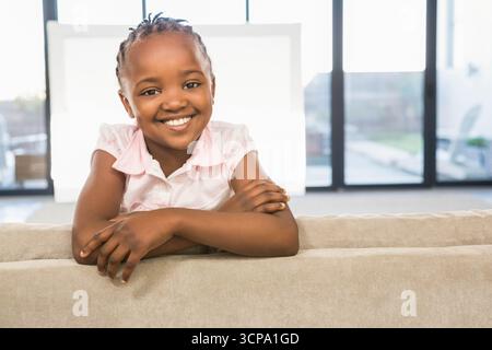 Fille afro-américaine penchée sur le canapé dos souriant à la caméra dans le salon à travers les panneaux de fenêtre Banque D'Images