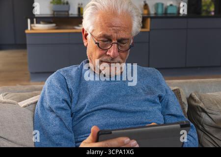 Homme senior portant un pull bleu assis sur un canapé avec des lunettes et utilisant une tablette dans le salon de la cuisine Banque D'Images