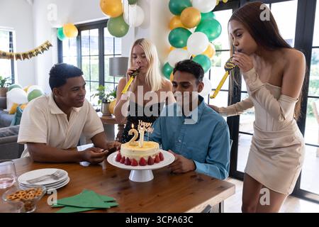 Divers amis se réunissant autour de la table à la maison célébrant gâteau d'anniversaire, ballons Banque D'Images