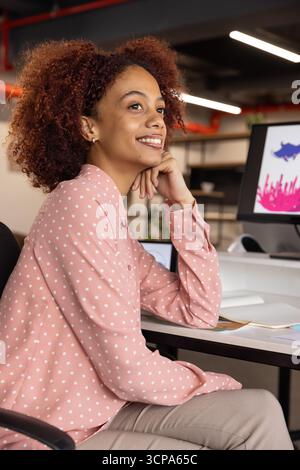 Femme afro-américaine assise au bureau dans le bureau, menton reposant sur la main, souriant à l'écran d'ordinateur Banque D'Images