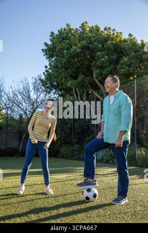 Couple asiatique debout dans la cour herbeuse avec le pied reposant sur le ballon de football à côté de la clôture à maillons de chaîne Banque D'Images