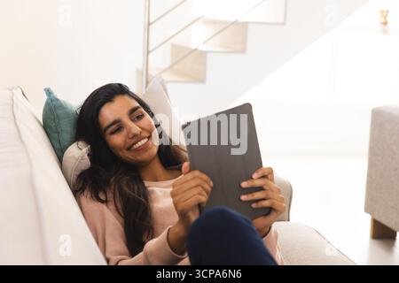 Femme asiatique souriante et tenant la tablette tout en s'inclinant sur le canapé avec coussin sarcelle dans le salon Banque D'Images