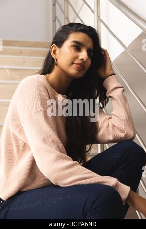 Femme sud-asiatique assise dans la cage d'escalier reposant la tête sur la main tout en regardant la rampe en acier inoxydable Banque D'Images