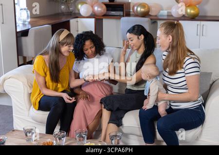 Diverses amies féminines assis sur le canapé examinant la main tout en tenant bébé à la maison, des ballons Banque D'Images