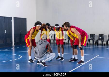 Équipe masculine diversifiée de basket-ball se blottissant autour de l'entraîneur sur le court, portant des maillots jaunes et rouges Banque D'Images