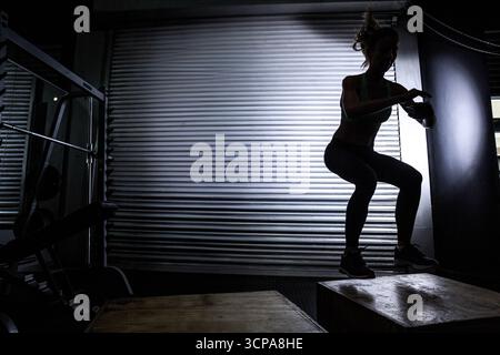 Athlète féminine adulte moyenne dans les vêtements de sport sautant sur la boîte plyo tenant kettlebell dans la salle de gym d'entrepôt Banque D'Images