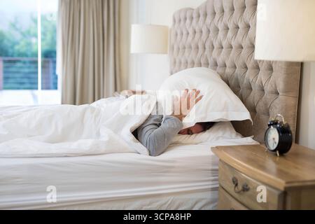 Homme couché sous couette blanche dans la chambre couvrant la tête avec oreiller sur le lit à côté de la lampe de table de nuit Banque D'Images