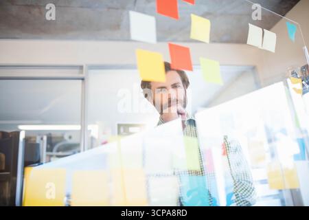 Homme barbu debout derrière un panneau de verre transparent examinant des notes adhésives dans un espace de travail ouvert Banque D'Images